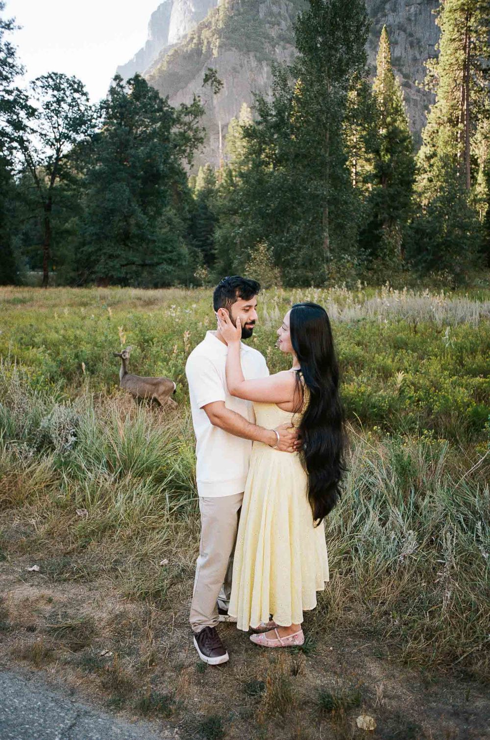 Couple embracing in Yosemite meadow with deer and waterfall backdrop, woman in yellow dress.