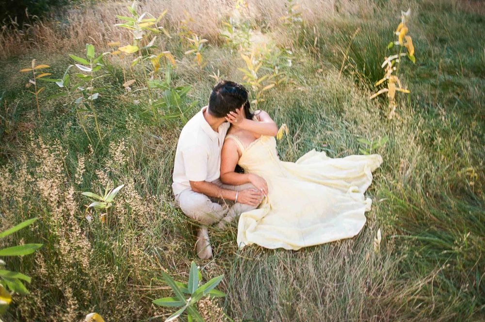 Couple sharing a romantic kiss in a lush green meadow, bride in flowing white gown seated in tall grass.