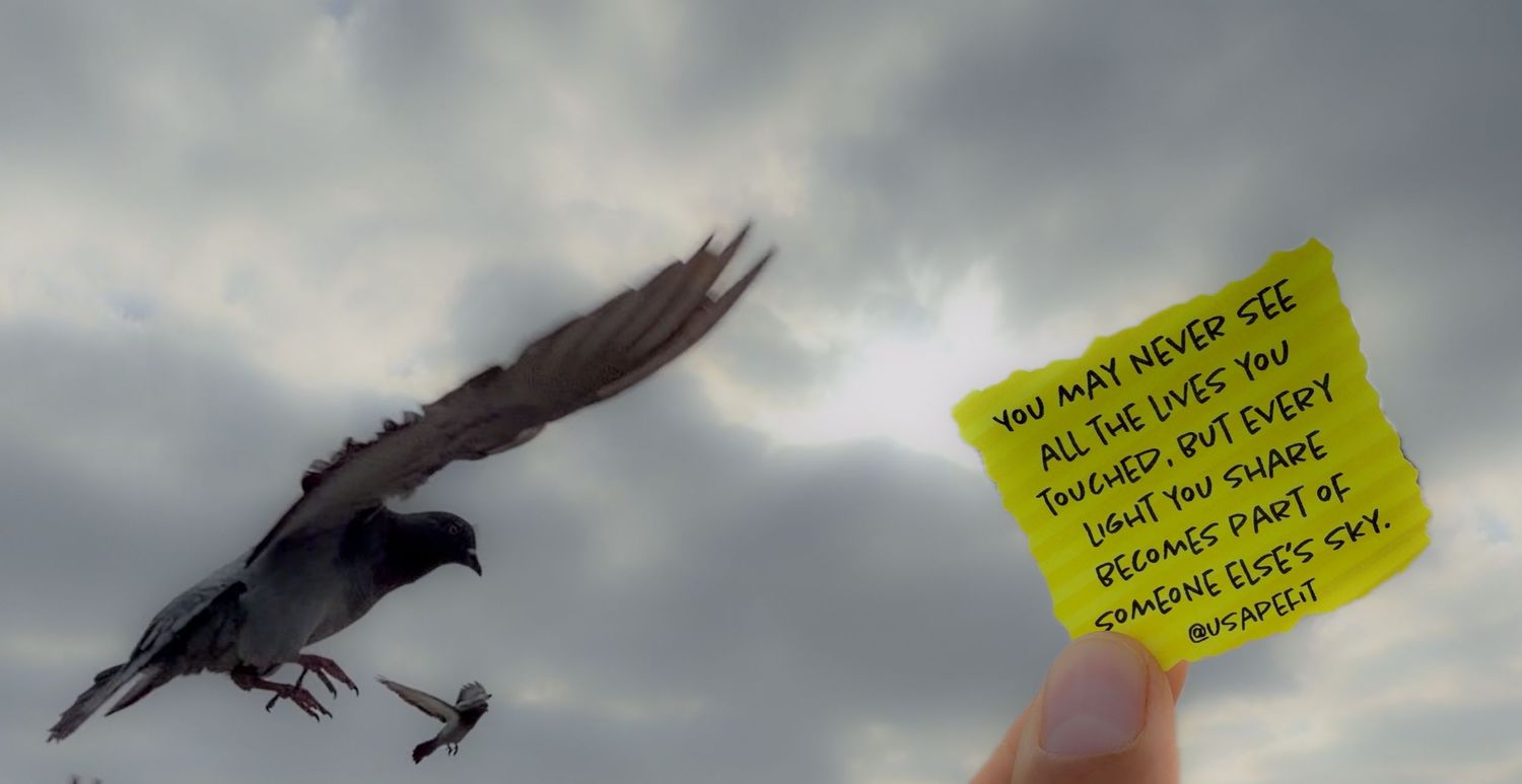 A crow in flight beside a hand holding a yellow sticky note with an inspirational quote against a cloudy sky.