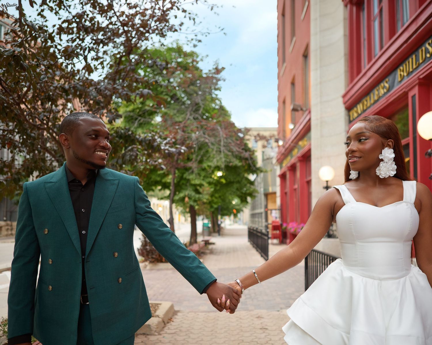 Couple holding hands on urban street, groom in green blazer, bride in white mini dress.