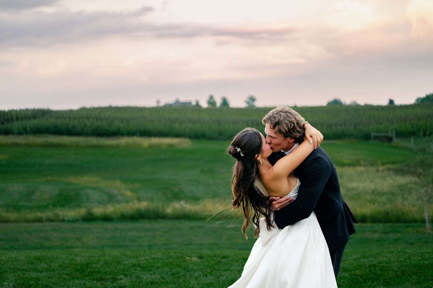 Bride and groom share romantic kiss on lush green farm field at sunset during outdoor wedding ceremony.
