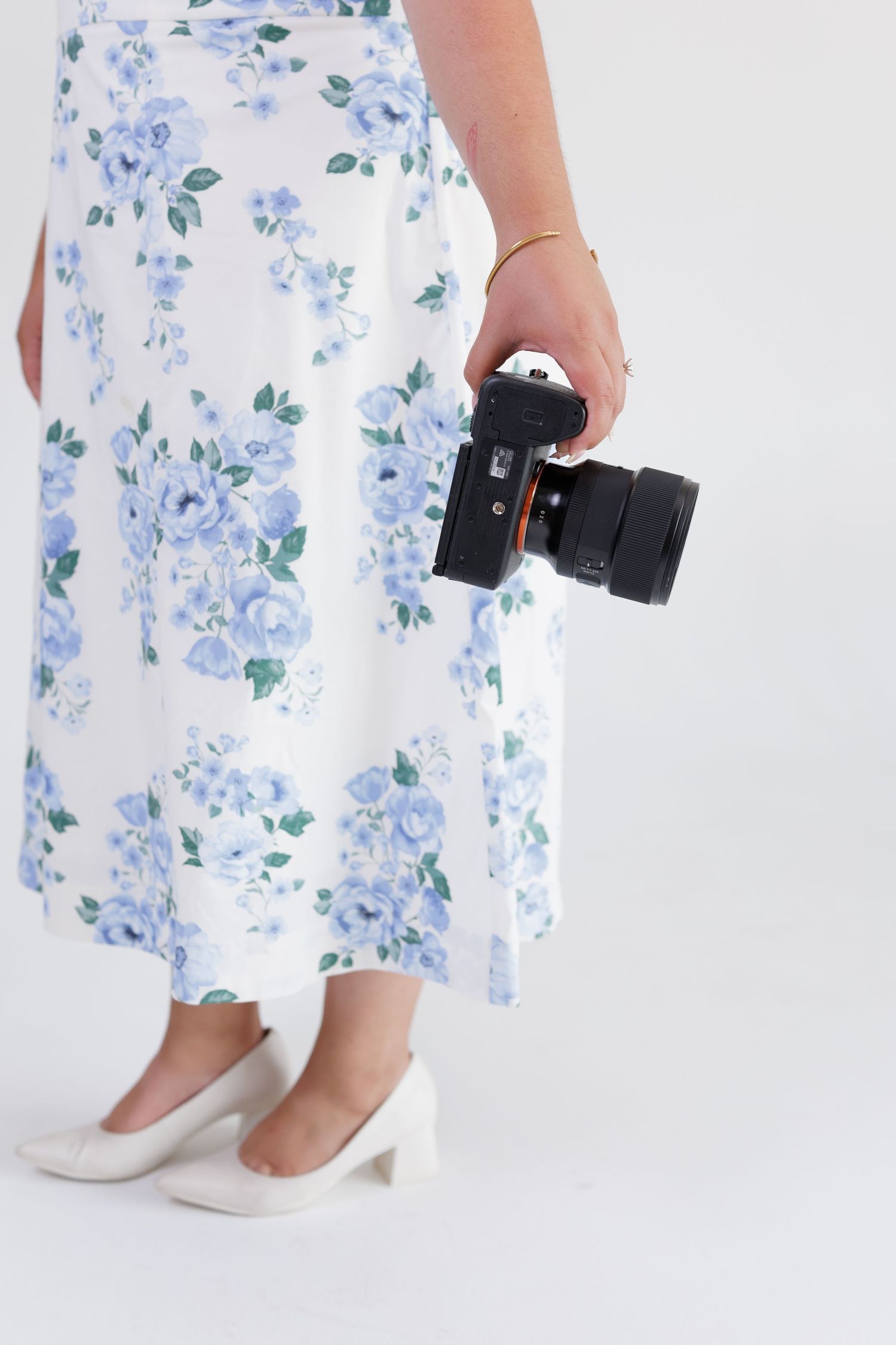 Woman in blue floral skirt and white heels holding a professional camera at her side.