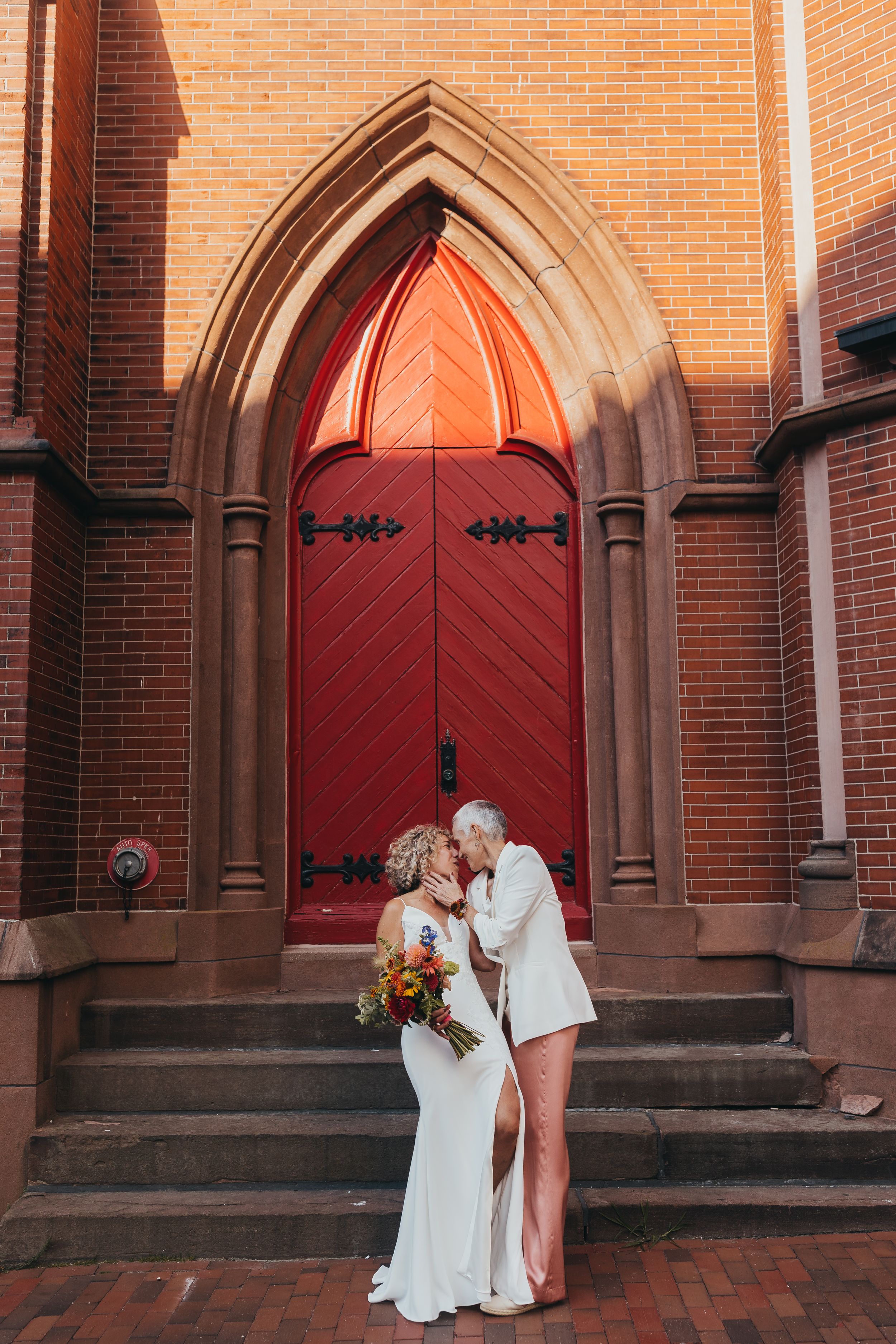 A couple stands in front of red church doors at a historic brick church with gothic arches.