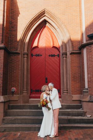 Couple poses for photos outside historic brick church with red doors at sunset.