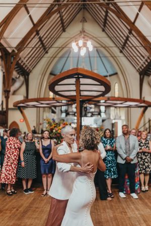 Wedding guests watching couple dance in rustic church hall with exposed beams and chandeliers.