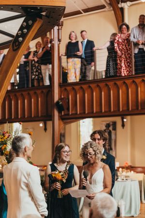 Wedding guests gathered on wooden balcony overlooking reception hall decorated with floral arrangements.