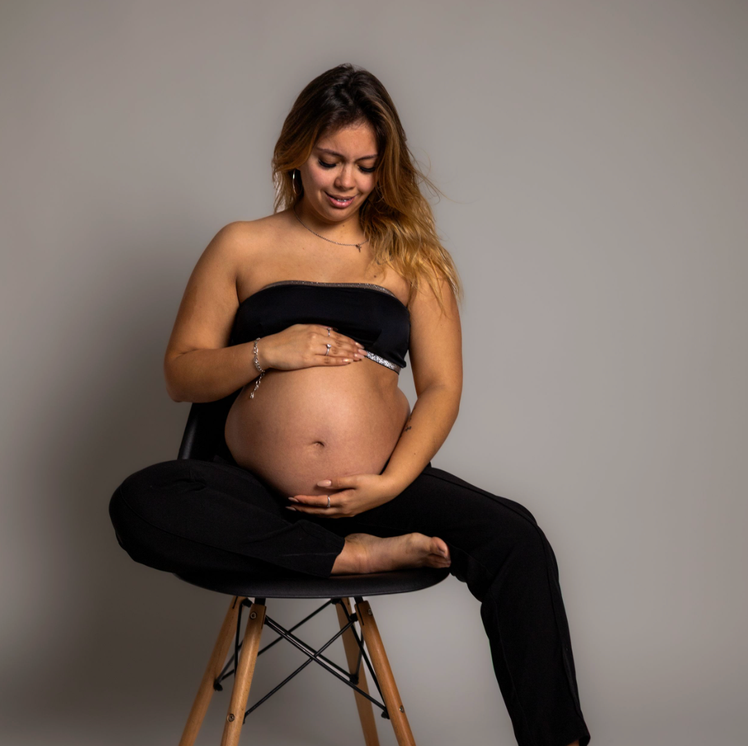 Pregnant woman in black outfit sitting on modern chair, cradling her baby bump in a professional maternity photo studio.