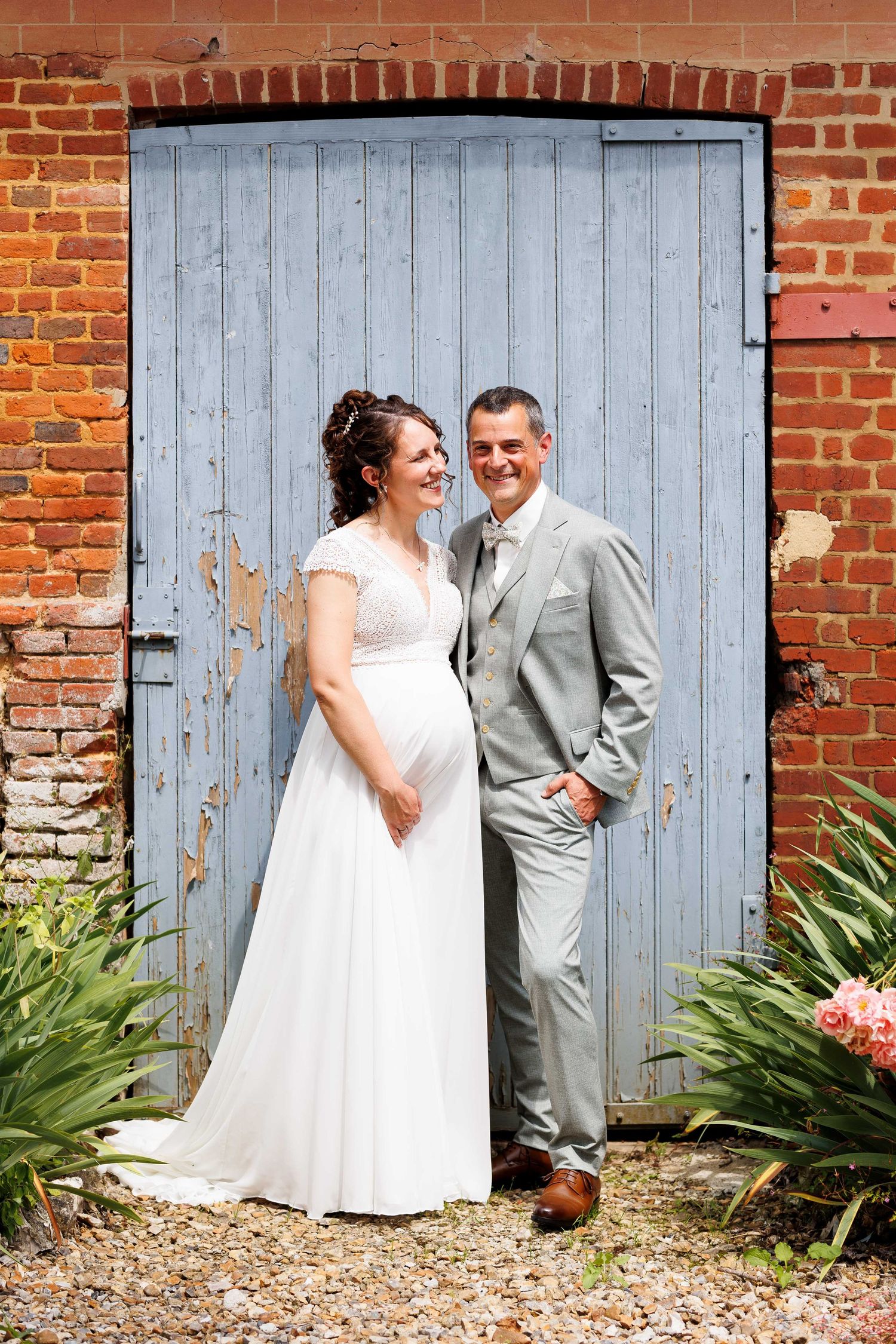 Pregnant bride in white gown and groom in gray suit pose before rustic blue door on their wedding day.