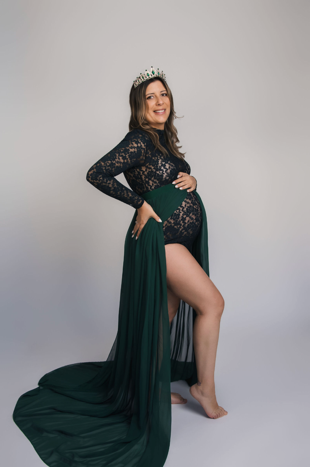 Pregnant woman in elegant green lace maternity gown with crown poses gracefully in studio photo shoot.