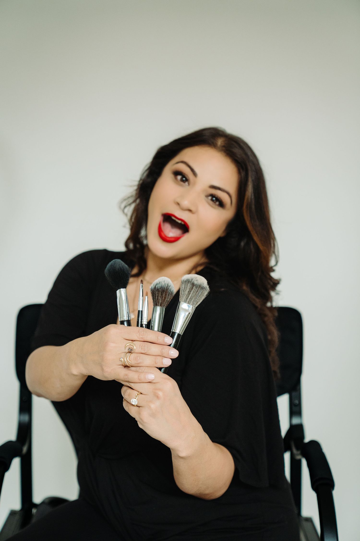 A makeup artist with red lips holds a collection of professional makeup brushes while sitting in a director's chair.