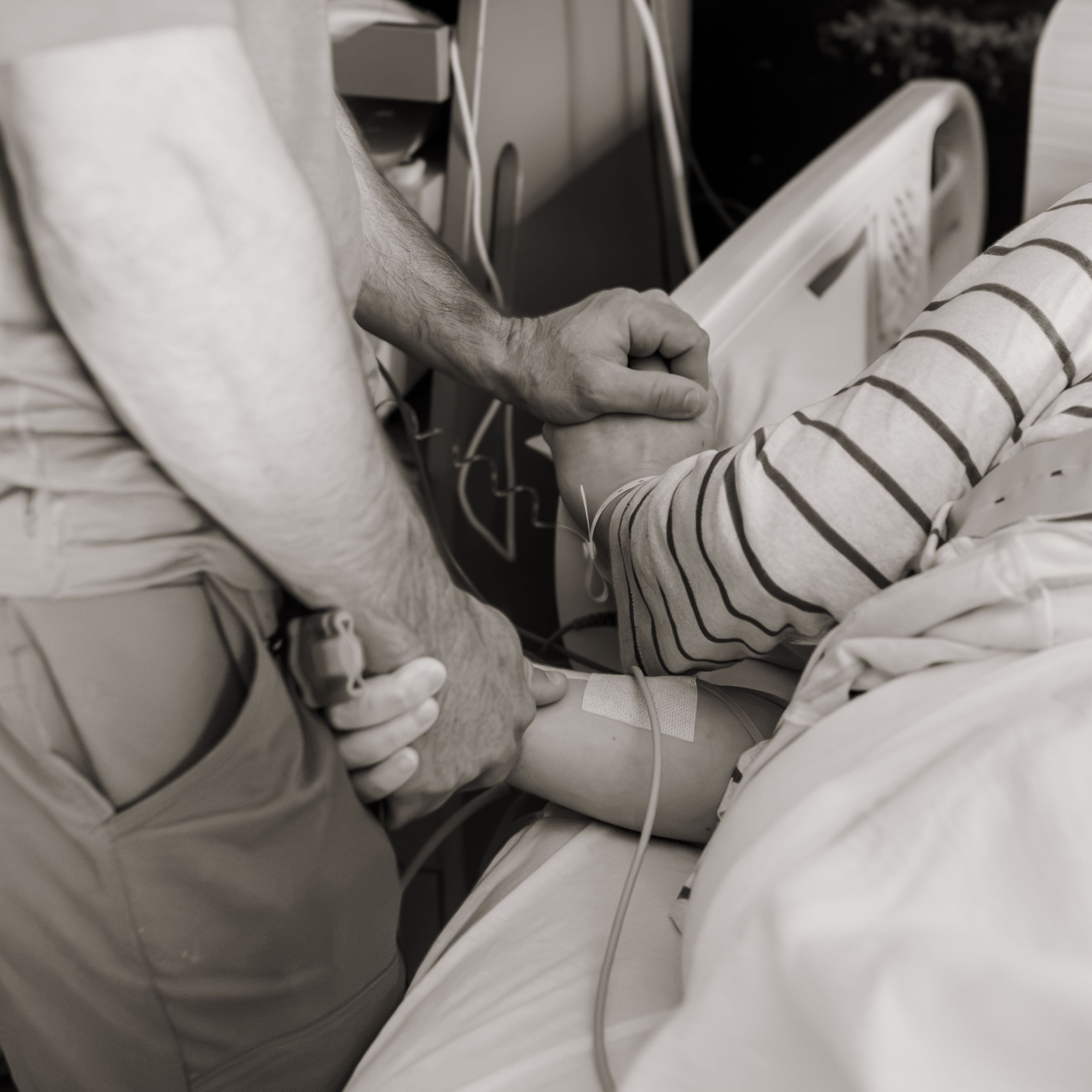 Black and white photo of a person holding a patient's hand in a hospital bed with medical equipment nearby.
