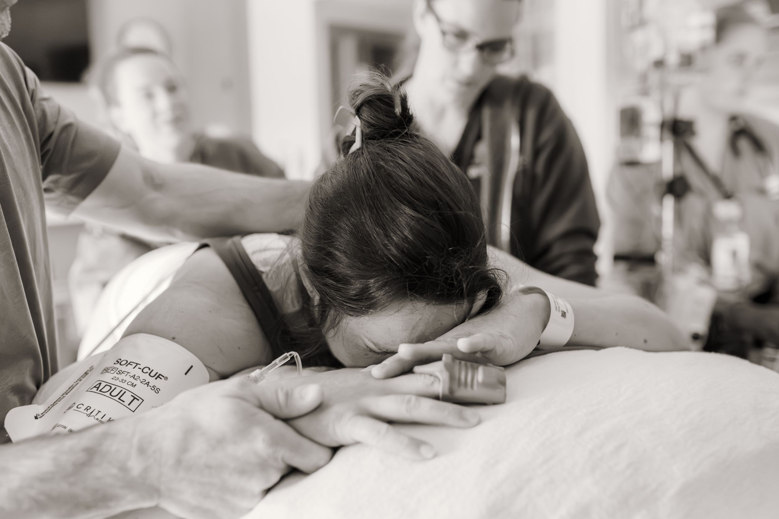 Black and white photo of a woman in labor leaning forward in pain, supported by medical staff in a hospital room.