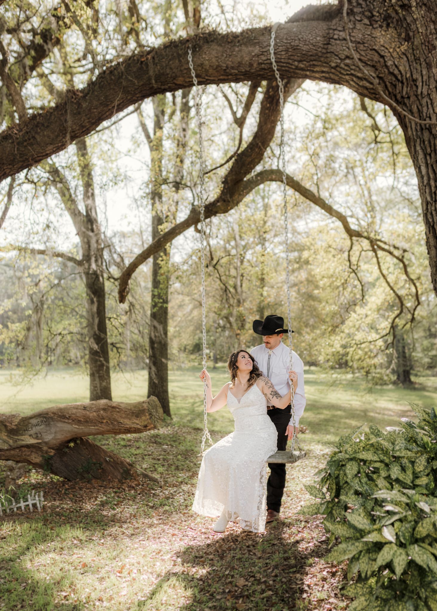 Bride sitting on a tree swing in a white dress while groom in black cowboy hat pushes her in a lush garden setting.