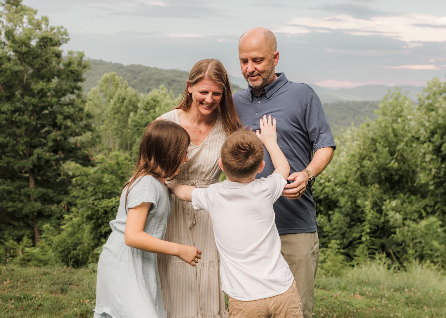 Family of four smiling outdoors with lush green trees and mountain views in the background.