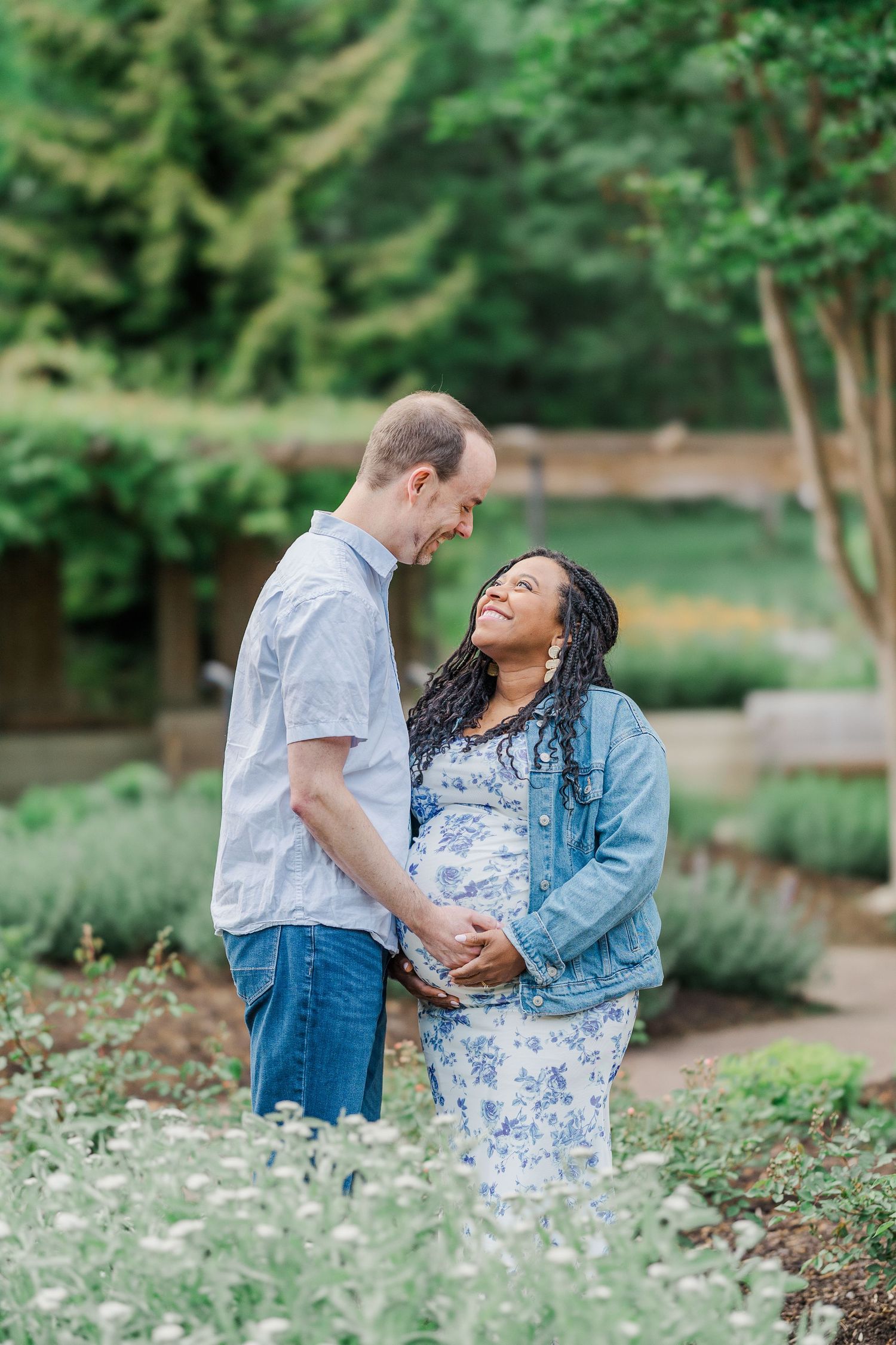 Interracial couple holding hands in a garden, smiling at each other during a maternity photoshoot outdoors.