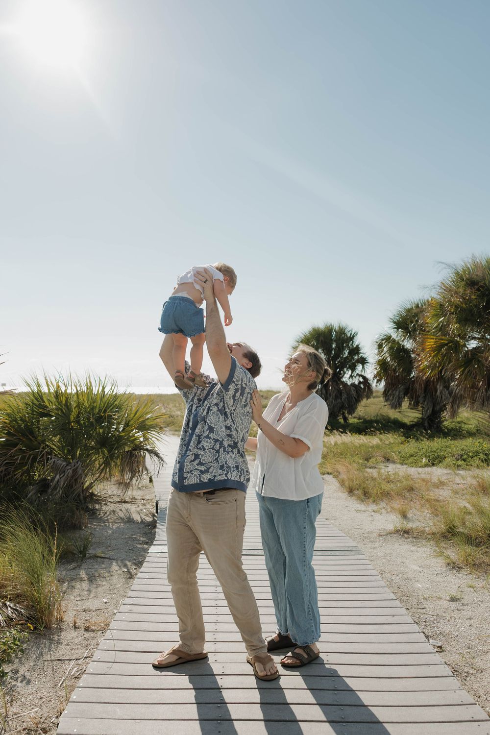 Family enjoying a sunny day on a beach boardwalk, with a man lifting a baby while a woman smiles nearby.