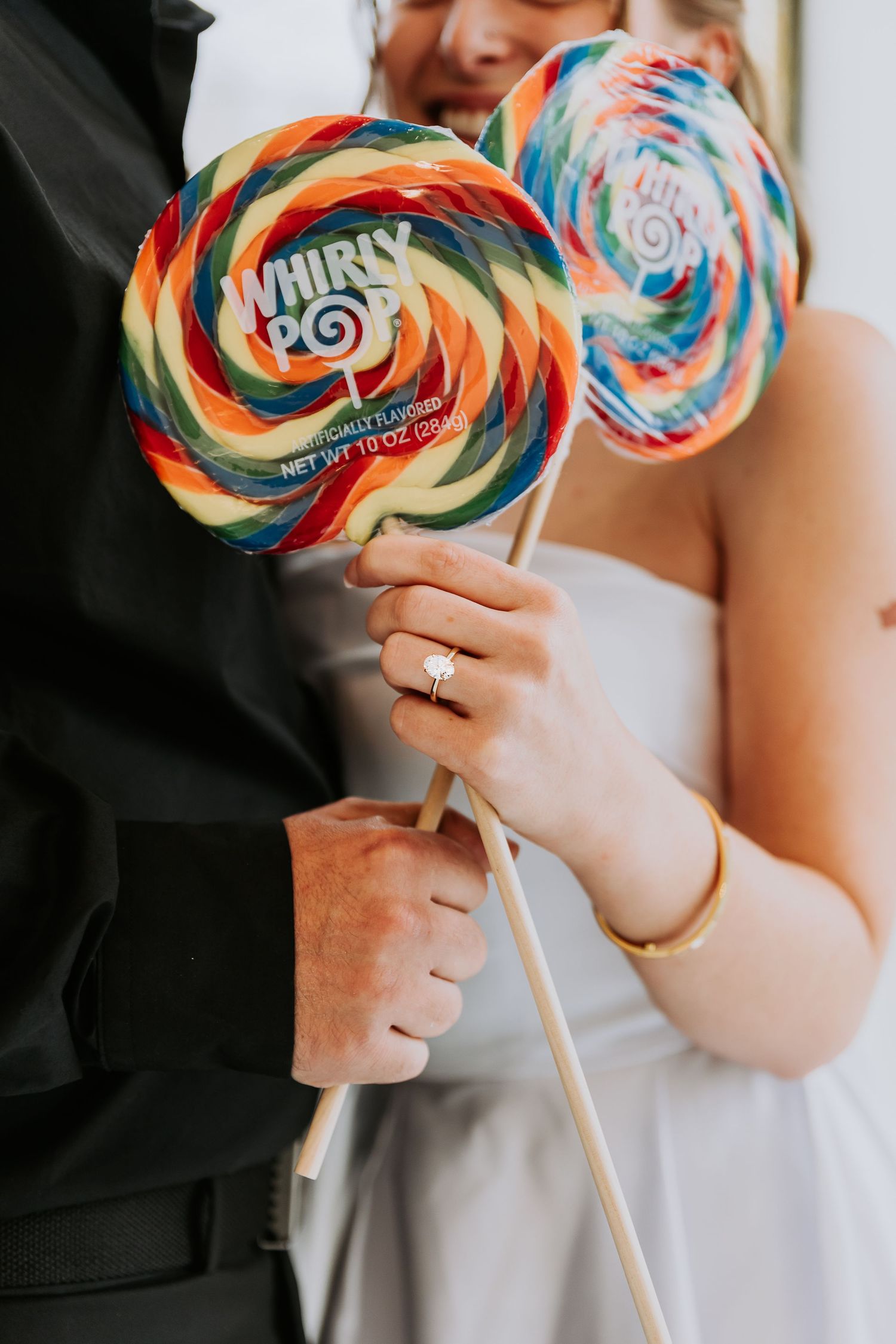Newlyweds holding colorful Whirly Pop lollipops, bride wearing engagement ring and gold bracelet in wedding attire.