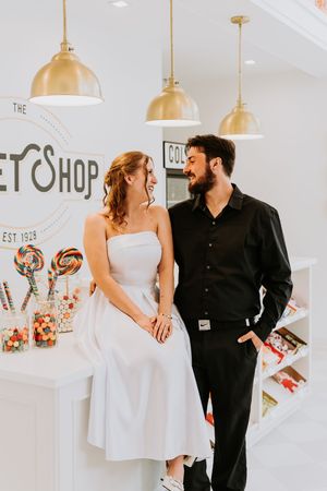 Couple smiling at each other in a charming candy shop with gold pendant lights and colorful lollipops on display.