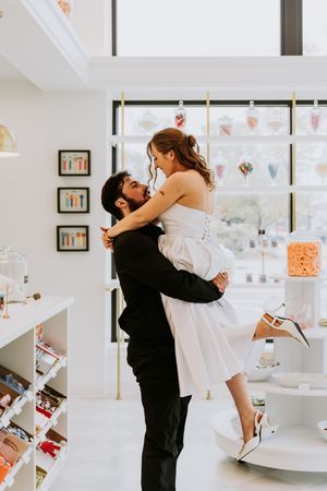 Groom lifting bride in white dress inside a bright candy shop, sharing a romantic moment.