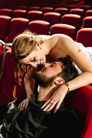 Couple sharing a romantic moment in red theater seats, woman leaning over bearded man smiling.