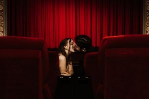 Couple sharing a romantic kiss in red velvet theater seats with dramatic crimson curtains backdrop.