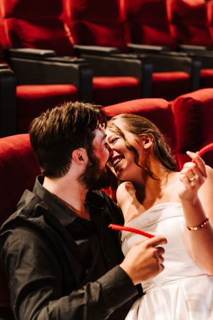 A couple shares a joyful moment in red theater seats, the woman in white laughing as the man leans in close.