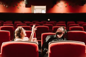 A man and woman tossing popcorn in an empty red theater, enjoying a playful moment among rows of seats.