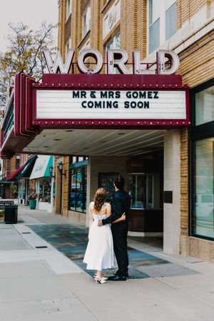 Couple stands under World Theatre marquee reading Mr & Mrs Gomez Coming Soon in a romantic wedding photo.