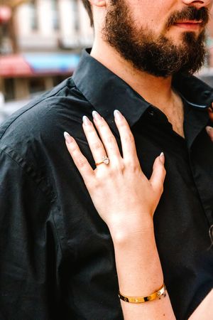 Woman's hand with diamond ring and gold bracelet resting on man's black shirt shoulder.