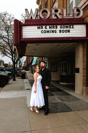 Couple poses under World Theatre marquee reading Mr & Mrs Gomez Coming Soon on a city sidewalk.
