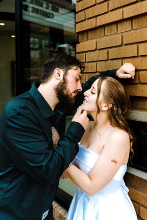 Romantic couple in wedding attire sharing an intimate moment against a brick wall outdoors.