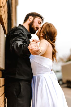 A couple in wedding attire holds hands and shares an intimate moment against a brick wall outdoors.