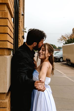 A couple in formal attire shares a romantic embrace against a brick wall, the woman smiling in a white strapless gown.