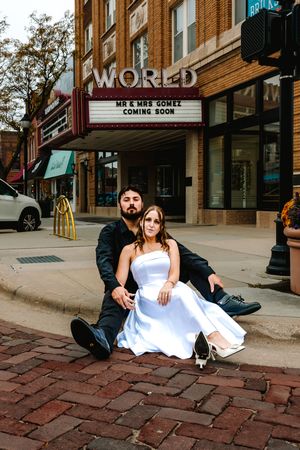 Newlyweds Mr. & Mrs. Gomez pose on brick steps in front of World Theater marquee, bride in white gown.