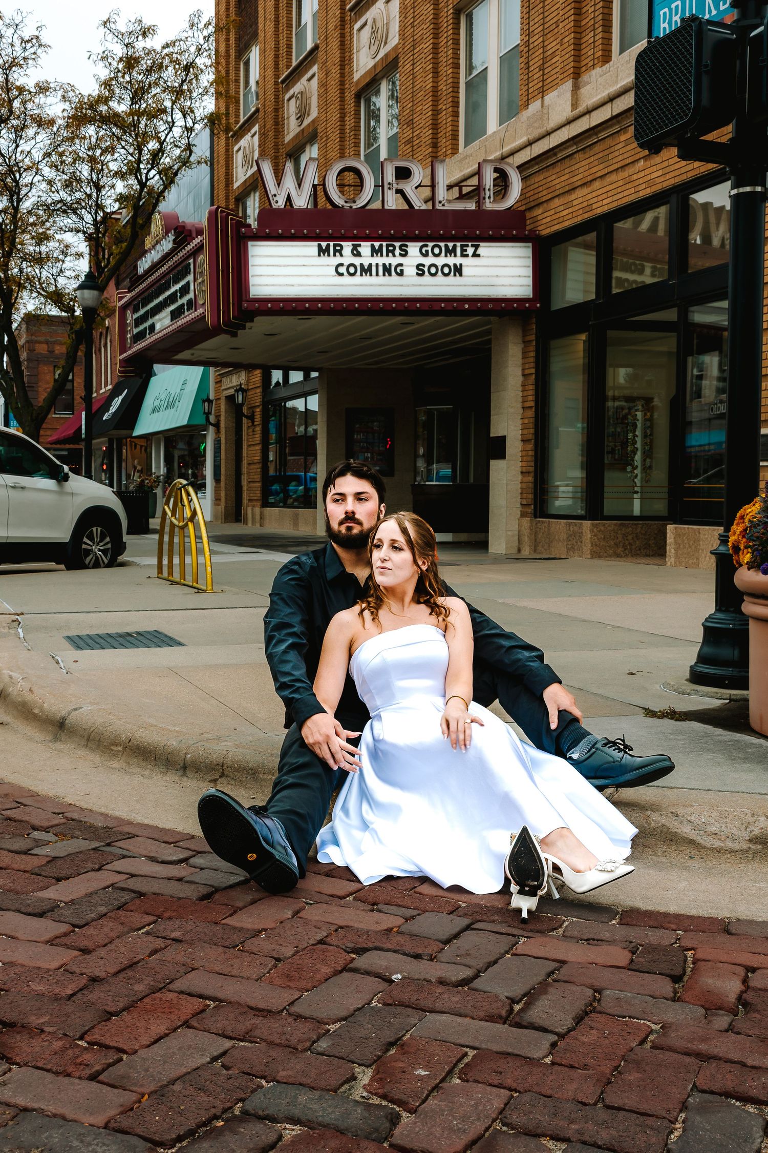 Newlyweds Mr & Mrs Gomez sit on brick sidewalk in wedding attire outside the World Theater marquee sign.