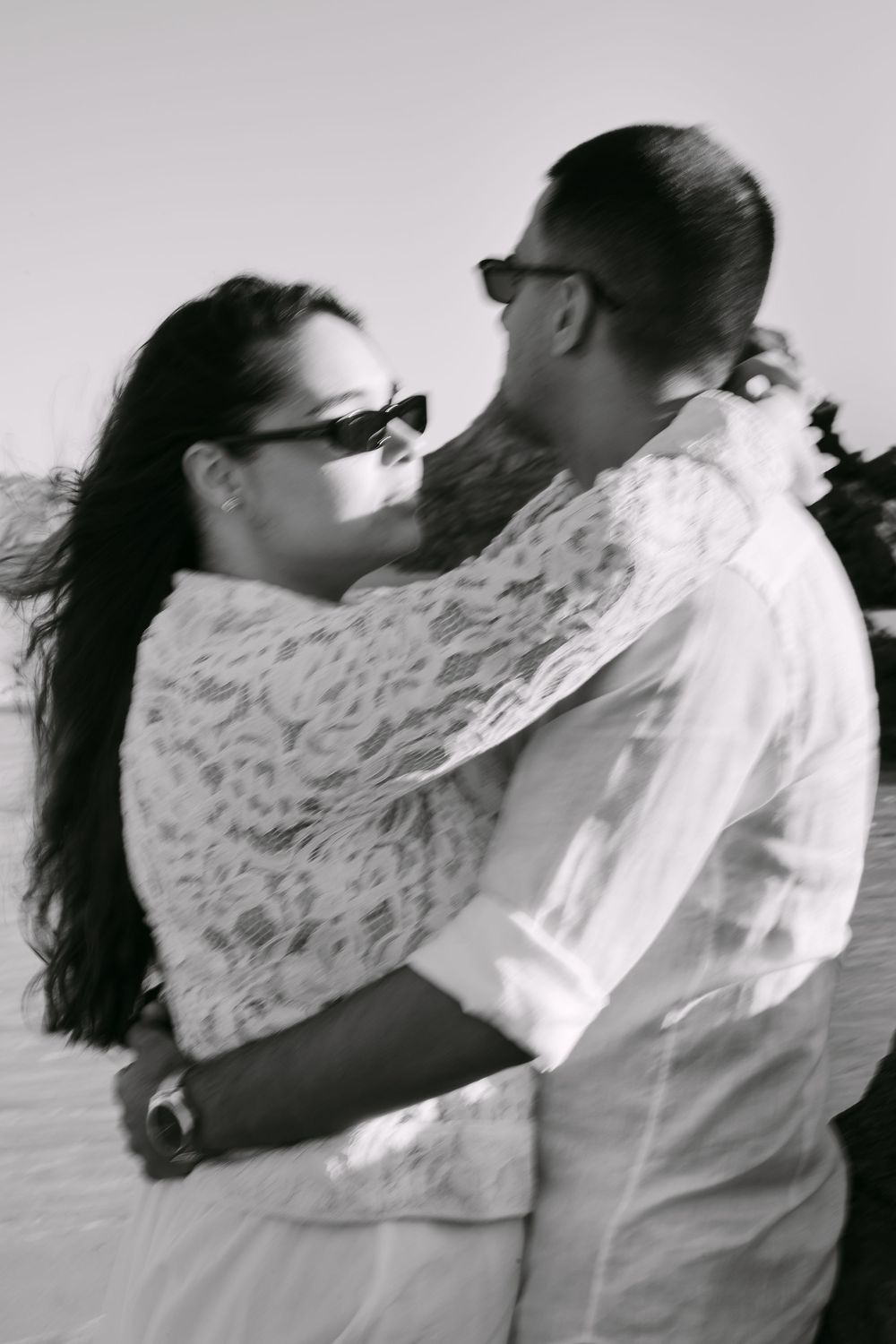 Black and white photo of a couple embracing outdoors, both wearing sunglasses, woman in lace dress, man in white shirt.