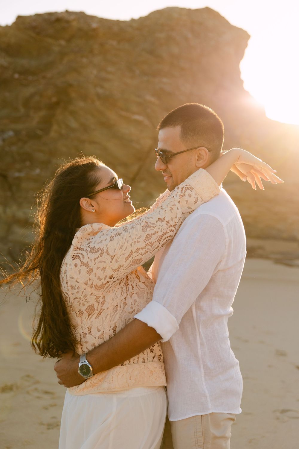 Couple embracing on a sandy beach at golden sunset, woman in lace top and man in white shirt, both wearing sunglasses.