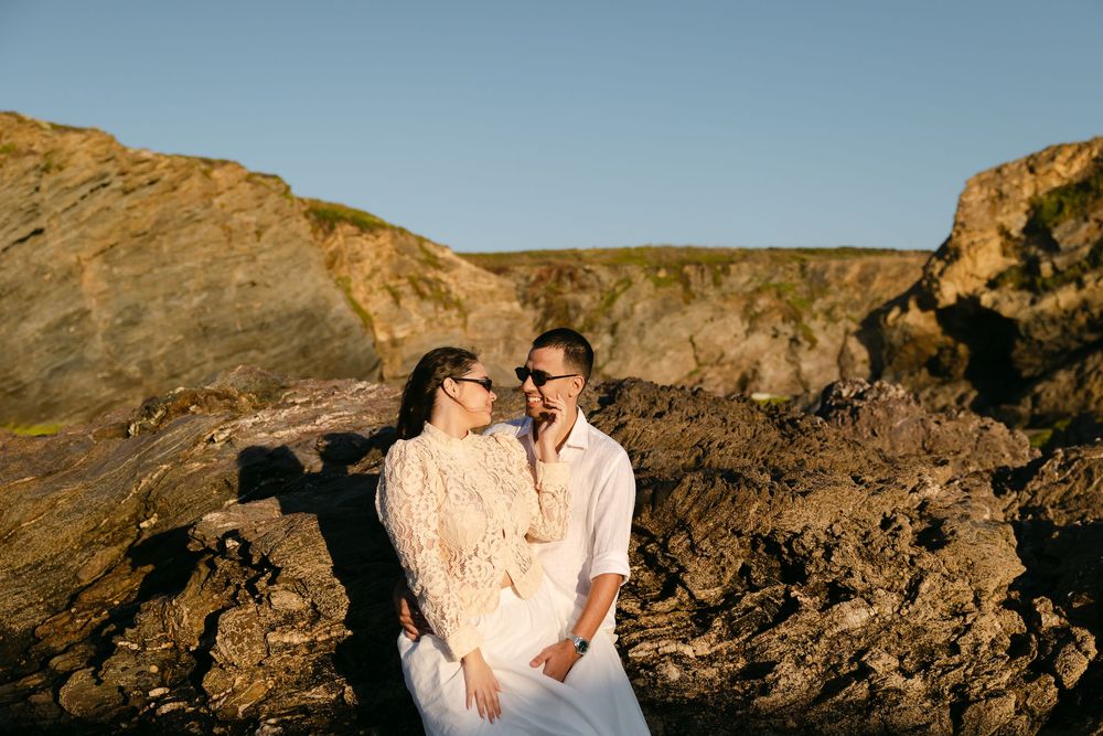 Couple in white outfits sharing a romantic moment among rocky coastal cliffs at golden hour sunset.
