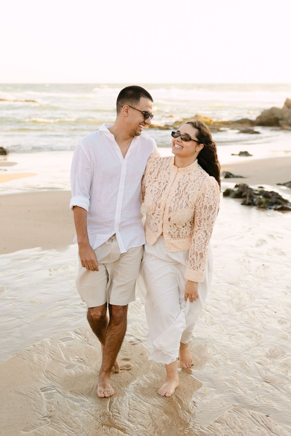 A couple walks barefoot on a sandy beach, smiling at each other, dressed in casual white and beige outfits with sunglasses.