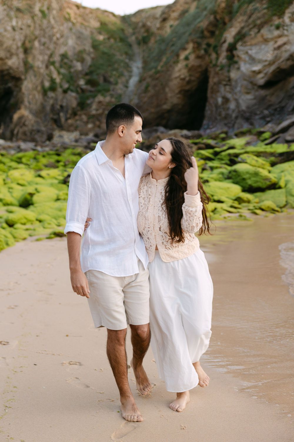 Couple walking barefoot on sandy beach near mossy rocks, man in white shirt and woman in flowing white skirt.