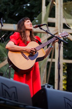 Woman in red dress playing acoustic guitar on outdoor stage with microphone stand.