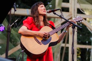 Woman in red dress playing acoustic guitar and singing into microphone on outdoor stage.
