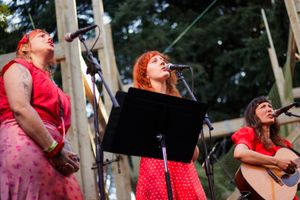 Three women in red perform on outdoor stage, two singing at microphones and one playing acoustic guitar.