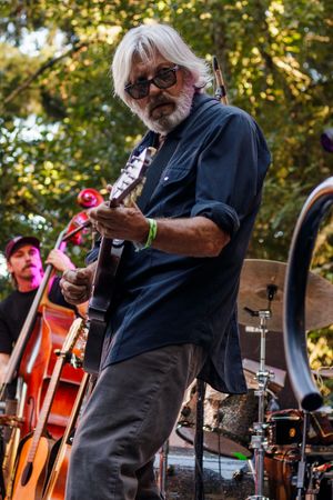 Elderly musician with white hair and beard plays electric guitar outdoors at a live music festival with trees in background.