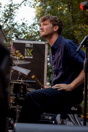 A young male drummer sits at a DW drum kit outdoors, playing near a Nexus equipment case surrounded by trees.