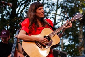 Woman in red dress playing acoustic guitar on stage outdoors at a live music performance.