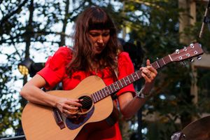 Woman in red dress playing acoustic guitar outdoors at a live music performance.