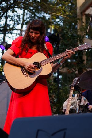 Woman in red dress playing acoustic guitar on outdoor stage with trees in background.
