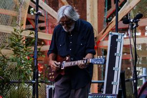 Elderly musician with white hair plays electric guitar on outdoor stage during live performance.