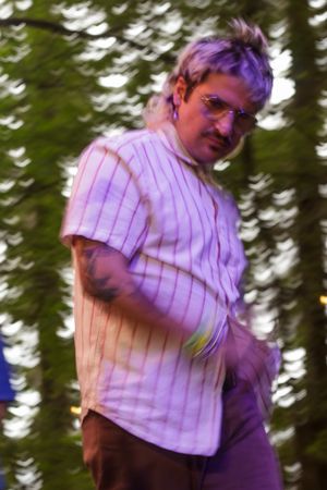 A tattooed man with purple hair and glasses wearing a striped shirt dances outdoors among green trees.